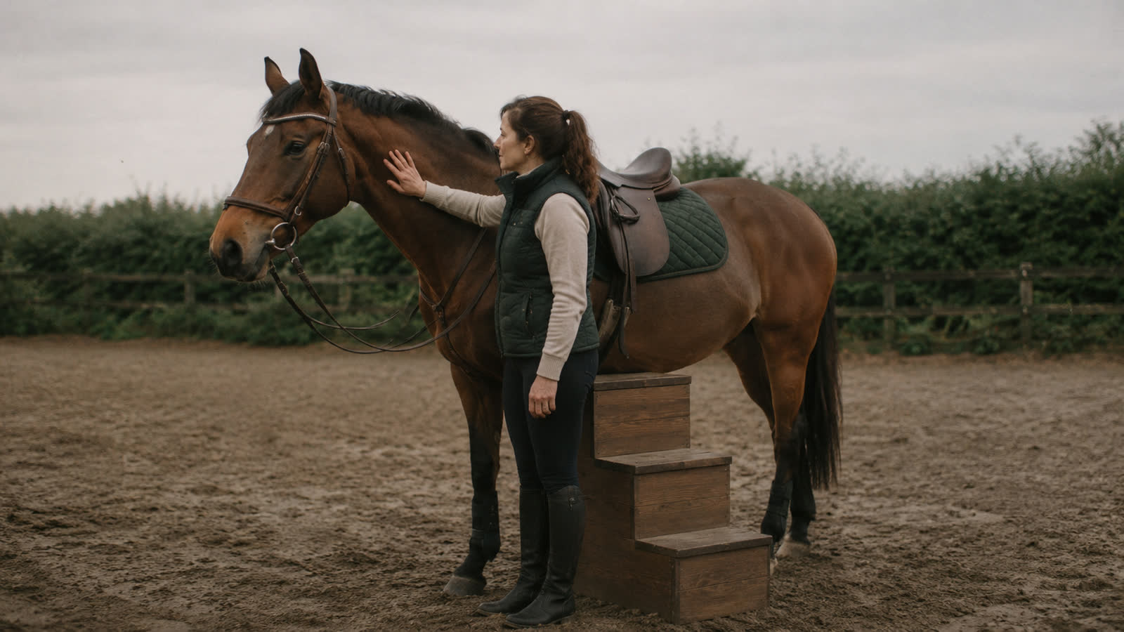 A rider standing at the mounting block with her hand on her horse's neck, tacked up, not mounting.